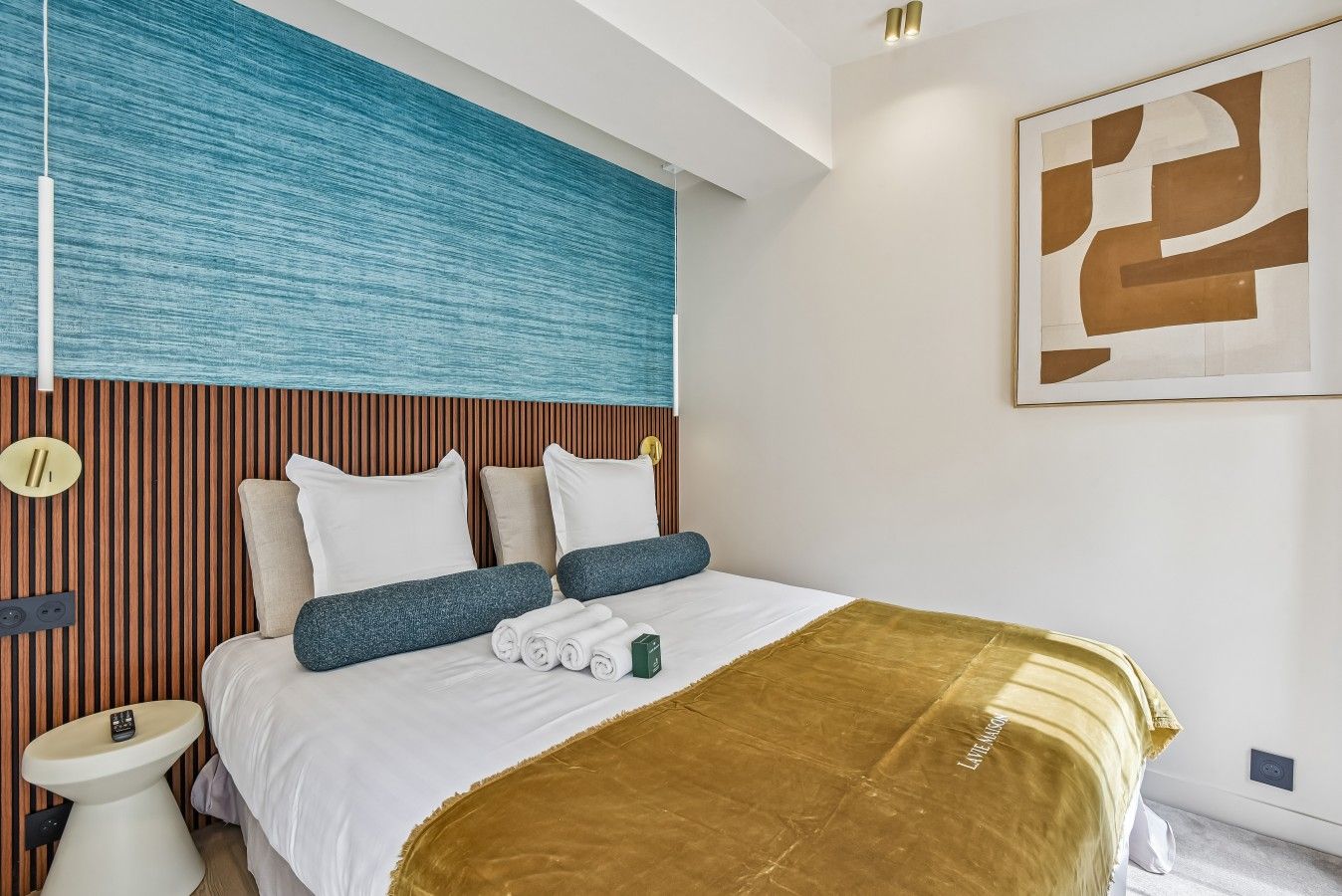 Ett modernt hotellrum med en stor säng, ett träfärgat sänggavel, en blå vägg med struktur och en stor, abstrakt målning.