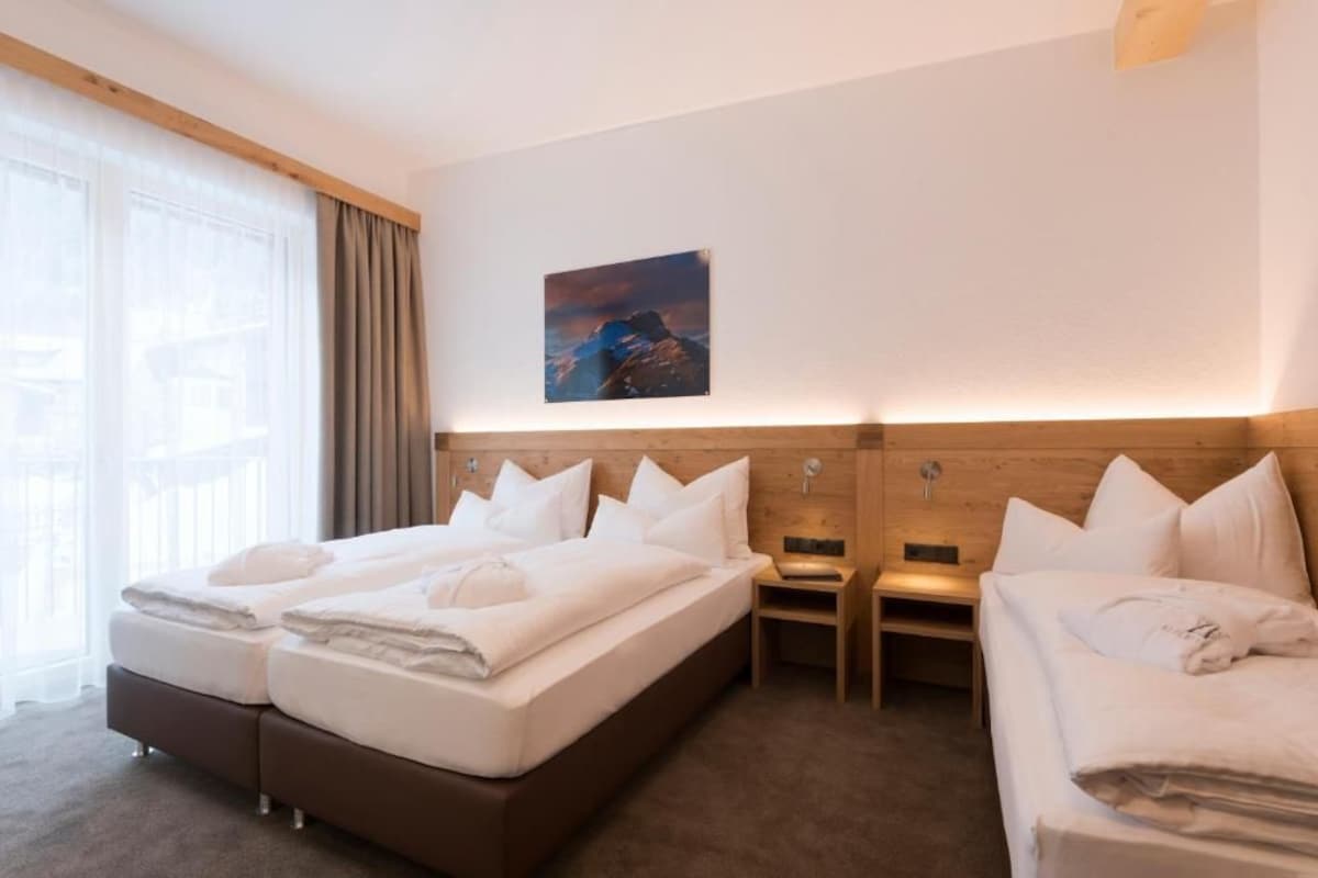 Ein Hotelzimmer mit einem großen Bett, einem kleineren Bett, einem Nachttisch, einem Wandbild und einem Fenster mit Vorhängen.