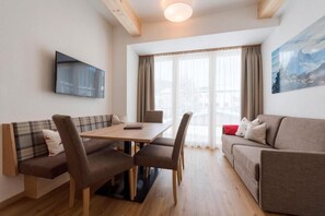 Uma sala de estar moderna com sofá, mesa de jantar com cadeiras, TV de tela plana na parede e uma grande janela com cortinas.
