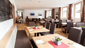 A hotel dining area with multiple tables and chairs, wooden flooring, and large windows with curtains. - Anthonys Peaks and Pillows St Anton (Sankt Anton am Arlberg)