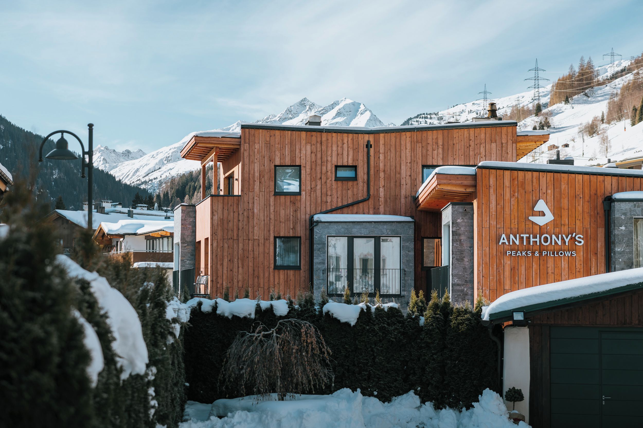 A modern wooden building with a sign reading 'ANTHONY'S PEAKS & PILLOWS' in a snowy mountainous area.