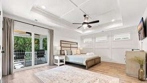 Ein Schlafzimmer mit einem Bett, einem Nachttisch, einem Schreibtisch, einem Sessel, einem Deckenventilator, einem Fernseher und Blick auf Grünfläche.