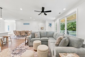 A modern living room with a grey sectional sofa, a dining area with wooden chairs, a ceiling fan, and a large window with a view of greenery. - Choose Happiness Updated 4 BR short golf cart ride Pawleys Pet Friendly (Pawleys Island)