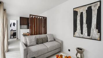 Ein Wohnzimmer mit einer Couch, einem Couchtisch mit Gebäck, einem Gemälde und einer Küche im Hintergrund.