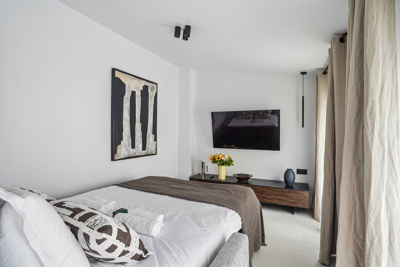 Ein modernes Schlafzimmer mit einem Bett, einem Flachbildfernseher, einem Konsolentisch mit Blumenvase und einem großen abstrakten Gemälde.