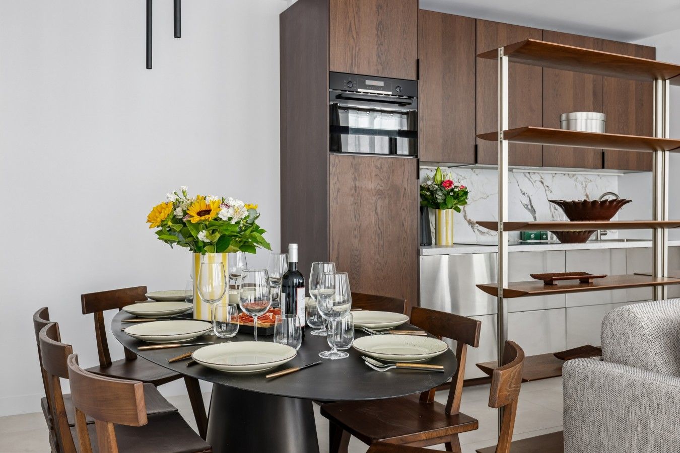 Un espace repas moderne avec une table ronde dressée pour six personnes, entourée de chaises en bois. Un vase rempli de fleurs jaunes est posé sur la table, ainsi que des verres à vin et une bouteille de vin.