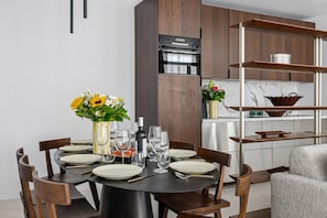 Un espace repas moderne avec une table ronde dressée pour six personnes, entourée de chaises en bois. Un vase rempli de fleurs jaunes est posé sur la table, ainsi que des verres à vin et une bouteille de vin.