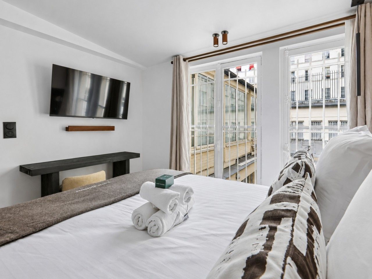 Une chambre d’hôtel avec un lit, un téléviseur à écran plat fixé au mur, une table de chevet et une fenêtre donnant sur la ville.