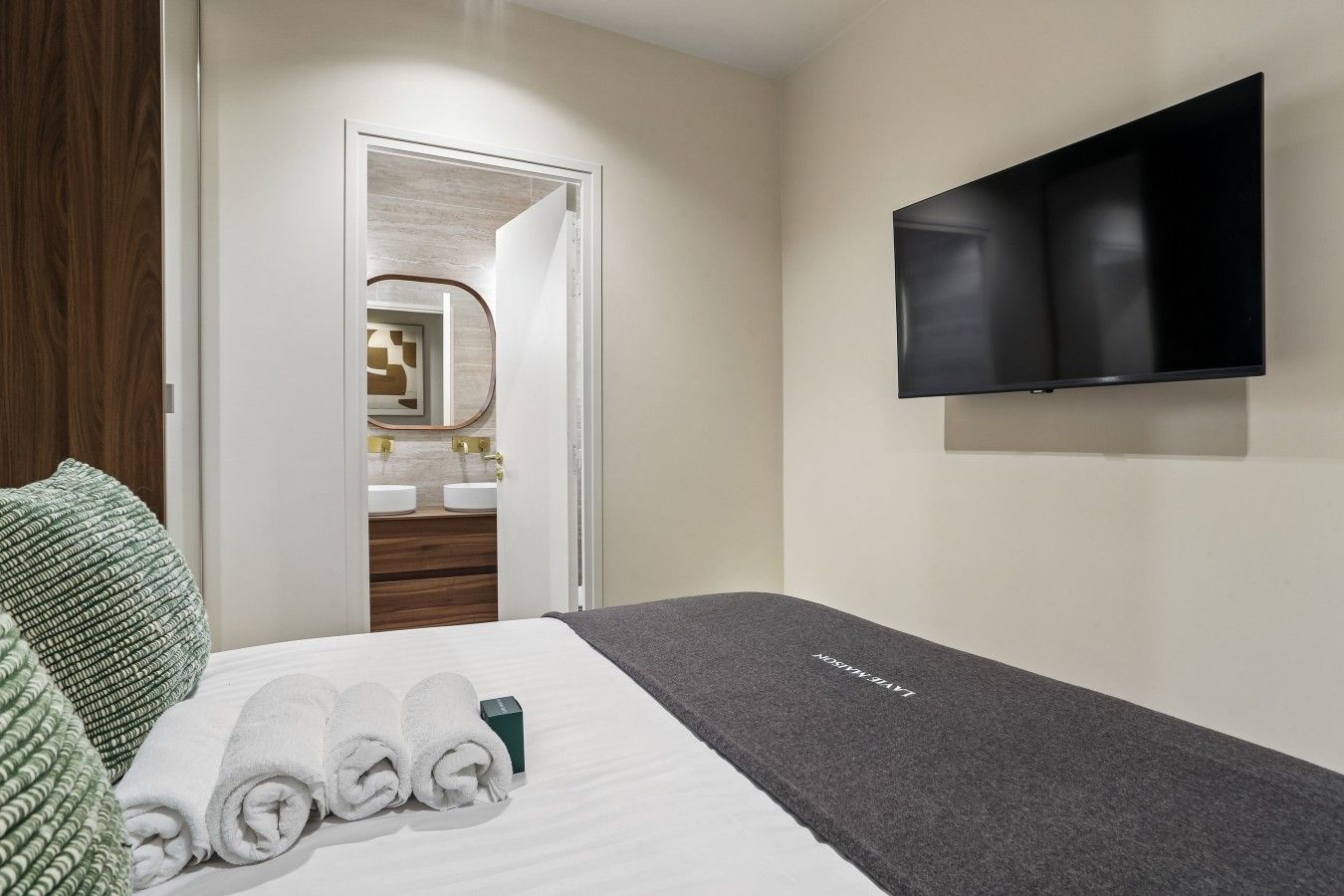 A hotel room with a bed, a TV, a bathroom with a mirror, and a green and white patterned pillow.