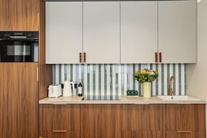 Una cocina moderna con armarios de madera, horno de acero inoxidable, encimera blanca, lavamanos con grifo dorado y un jarrón con flores.