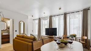 Ein modernes Wohnzimmer mit Sofa, Esstisch, Stühlen, Fernseher und einem großen Fenster mit Vorhängen.