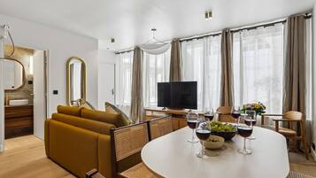 Een moderne woonkamer met een bank, een eettafel en stoelen, een televisie en een groot raam met gordijnen.