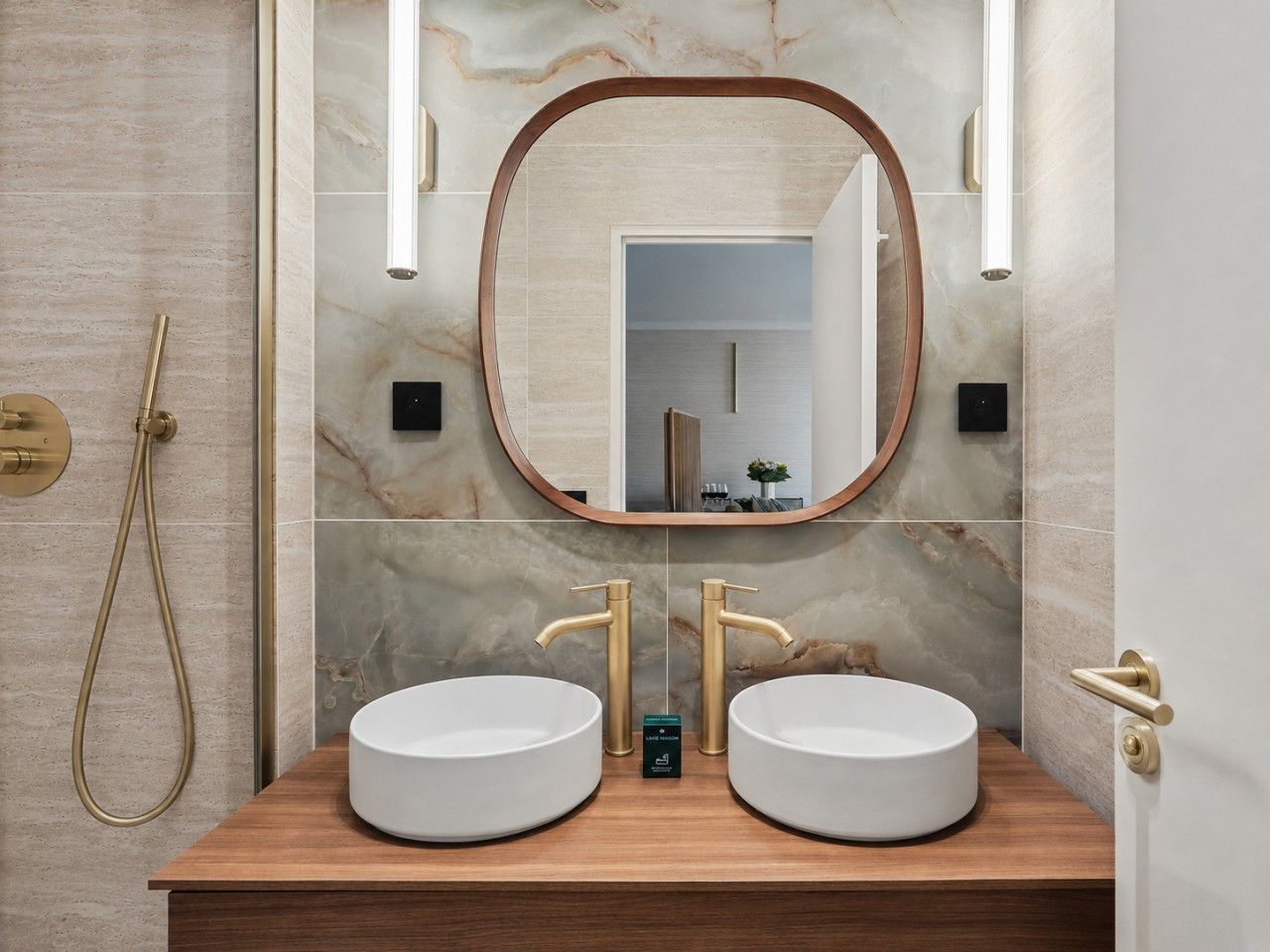 A bathroom with two round white sinks, a wooden vanity, a gold showerhead, and a marble wall.