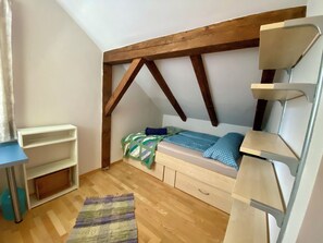 A small, cozy room with a sloped ceiling, wooden beams, a bed with a blue and green patterned blanket, a white shelf unit, and a wooden floor. - Bergulme Vacation Rental with a View of the Tyrolean Mountains (Mutters)