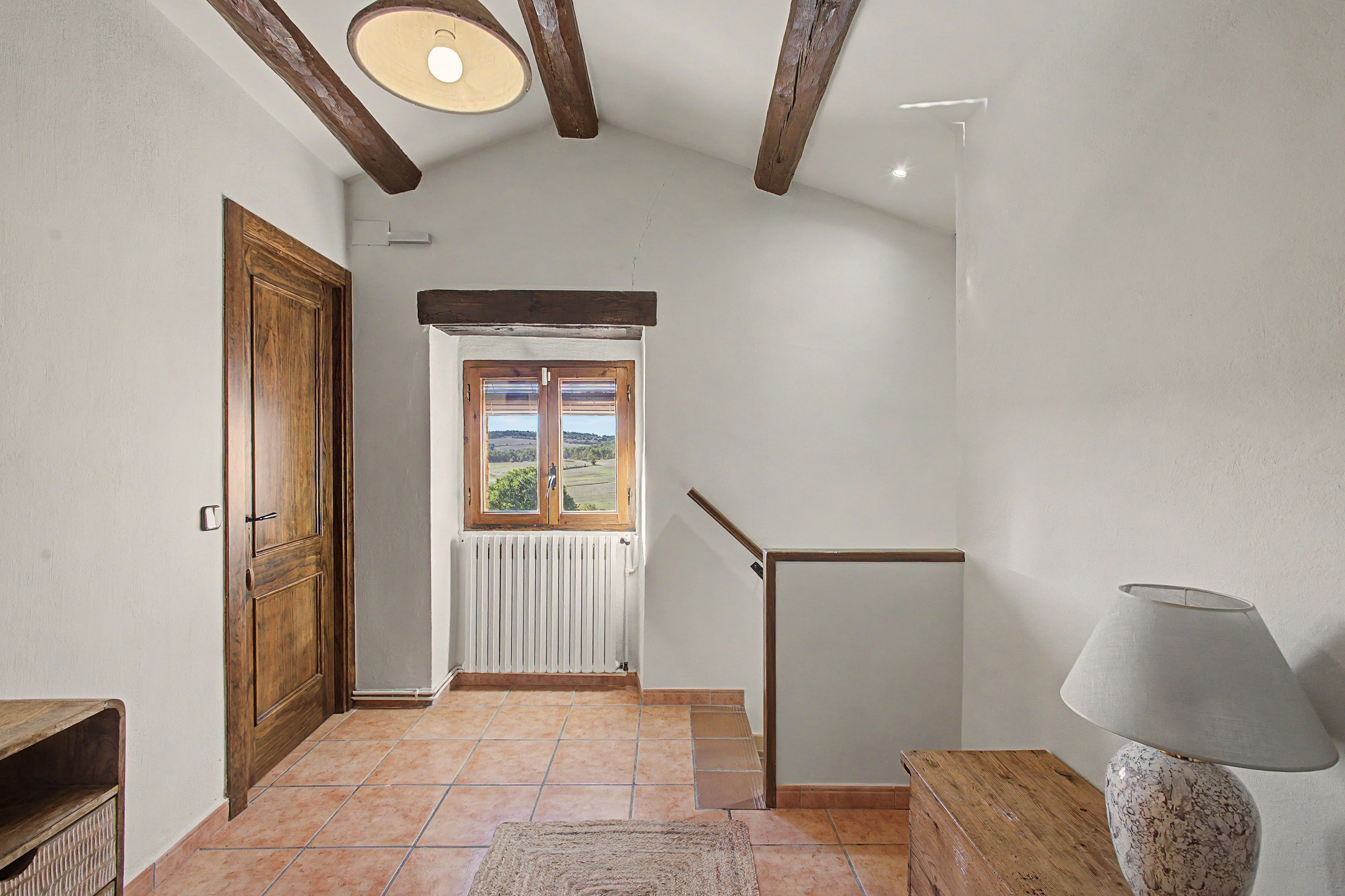 Un pasillo con una puerta de madera, una ventana con vistas, un radiador y una lámpara sobre una mesa.