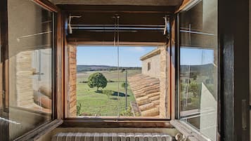 Habitació amb una finestra gran que ofereix vista a un paisatge rural, amb un arbre i un edifici.