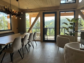 Uma área de refeições com mesa e cadeiras de madeira, um lustre e vista para o exterior.