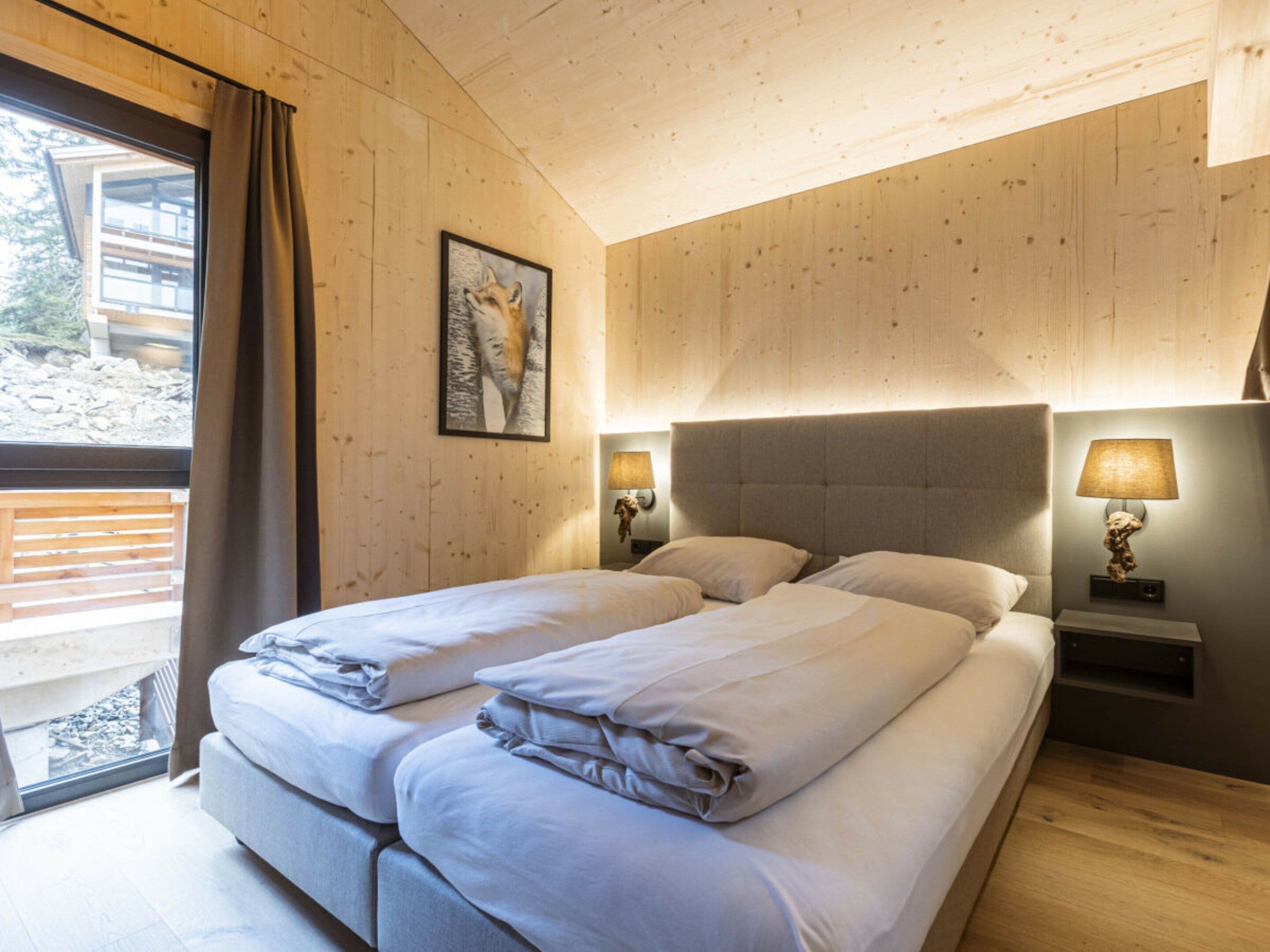 Een tweepersoonsbed met hoofdbord, twee nachtkastjes met lampen, een ingelijst schilderij aan de muur, en een raam met gordijnen.