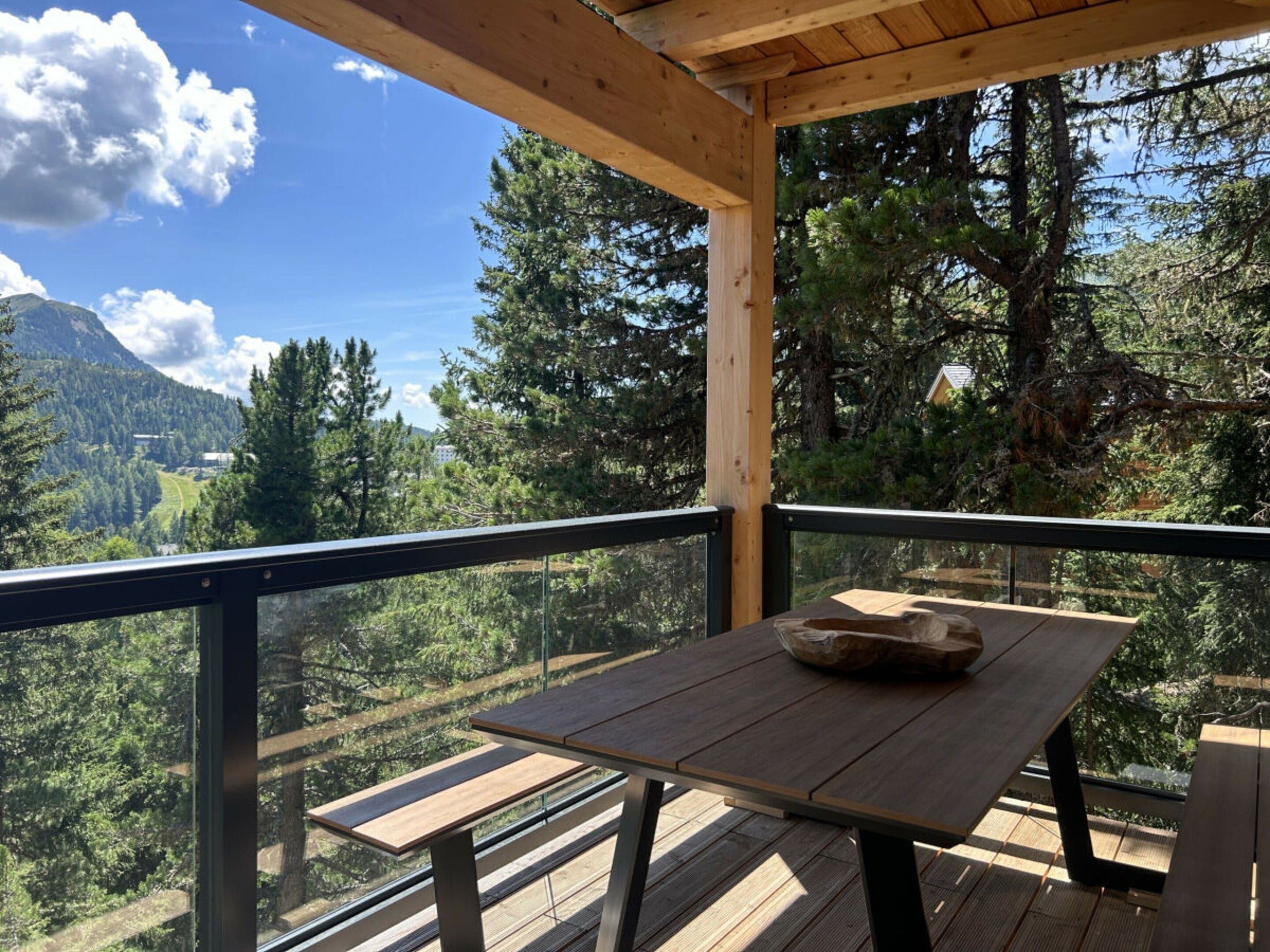 Een houten terras met een tafel en banken, met uitzicht op een bebost dal en bergen.