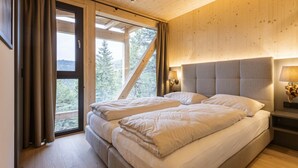 Ein Schlafzimmer mit einem großen Bett, einem Nachttisch, einer Lampe und Blick auf Bäume durch ein Fenster.