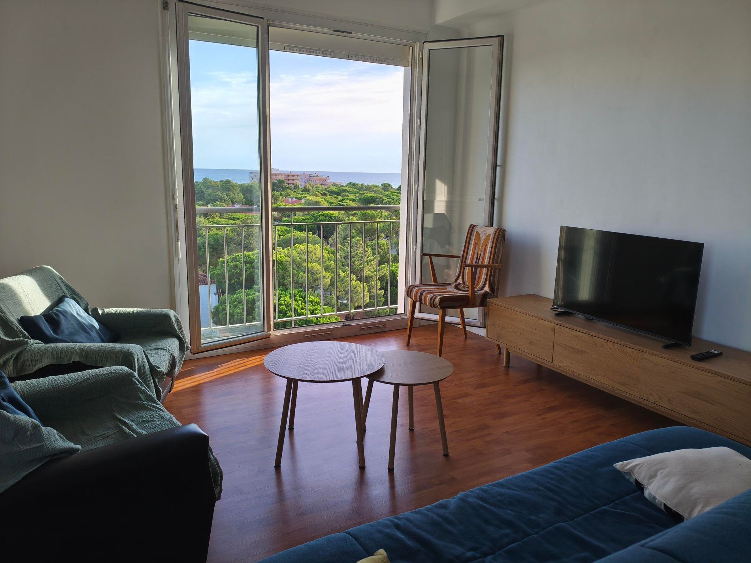 Sala de estar com sofá, televisão, armário de madeira e varanda com vista para áreas verdes e edifícios.