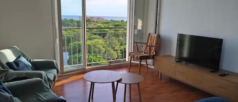 A living room with a sofa, a TV, a wooden cabinet, and a balcony with a view of greenery and buildings.