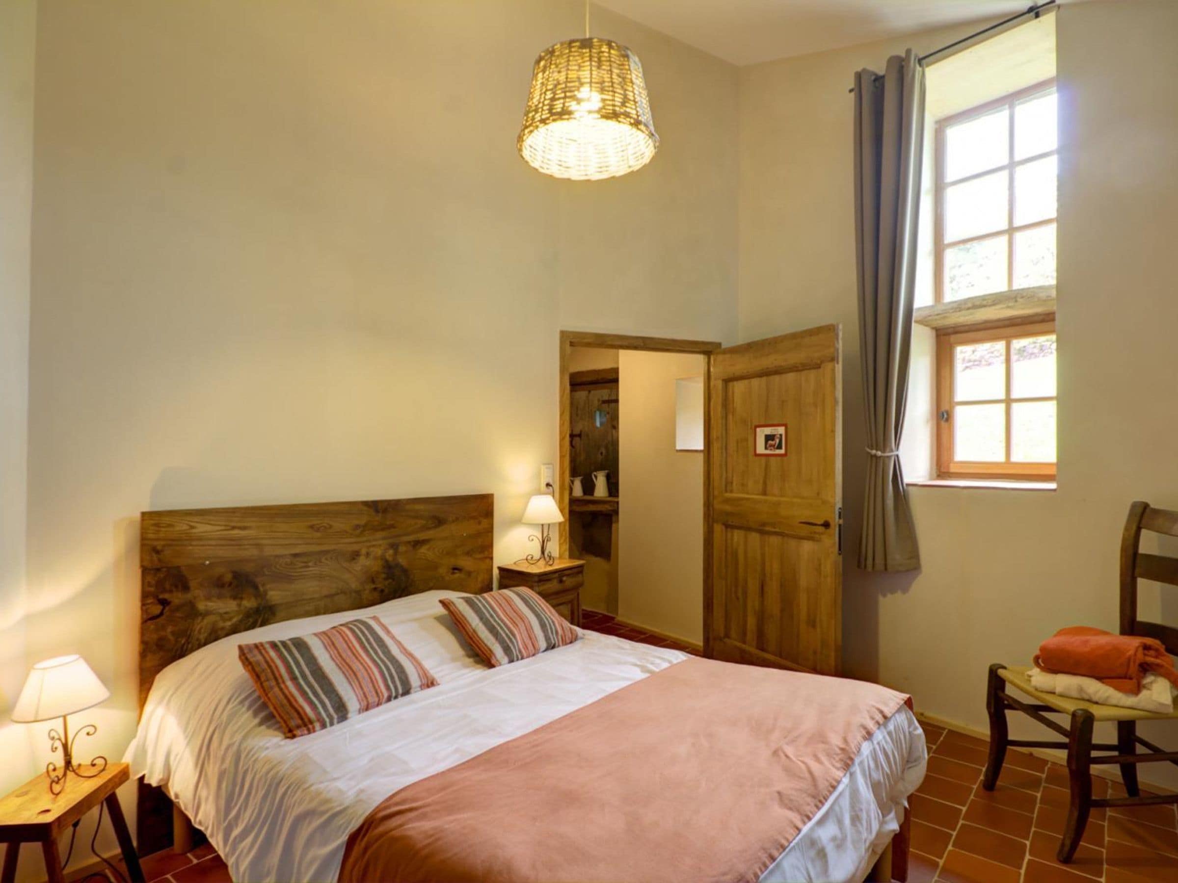 A bedroom with a wooden headboard, a bed with striped pillows, a bedside lamp, a window with curtains, and a chair with a towel.