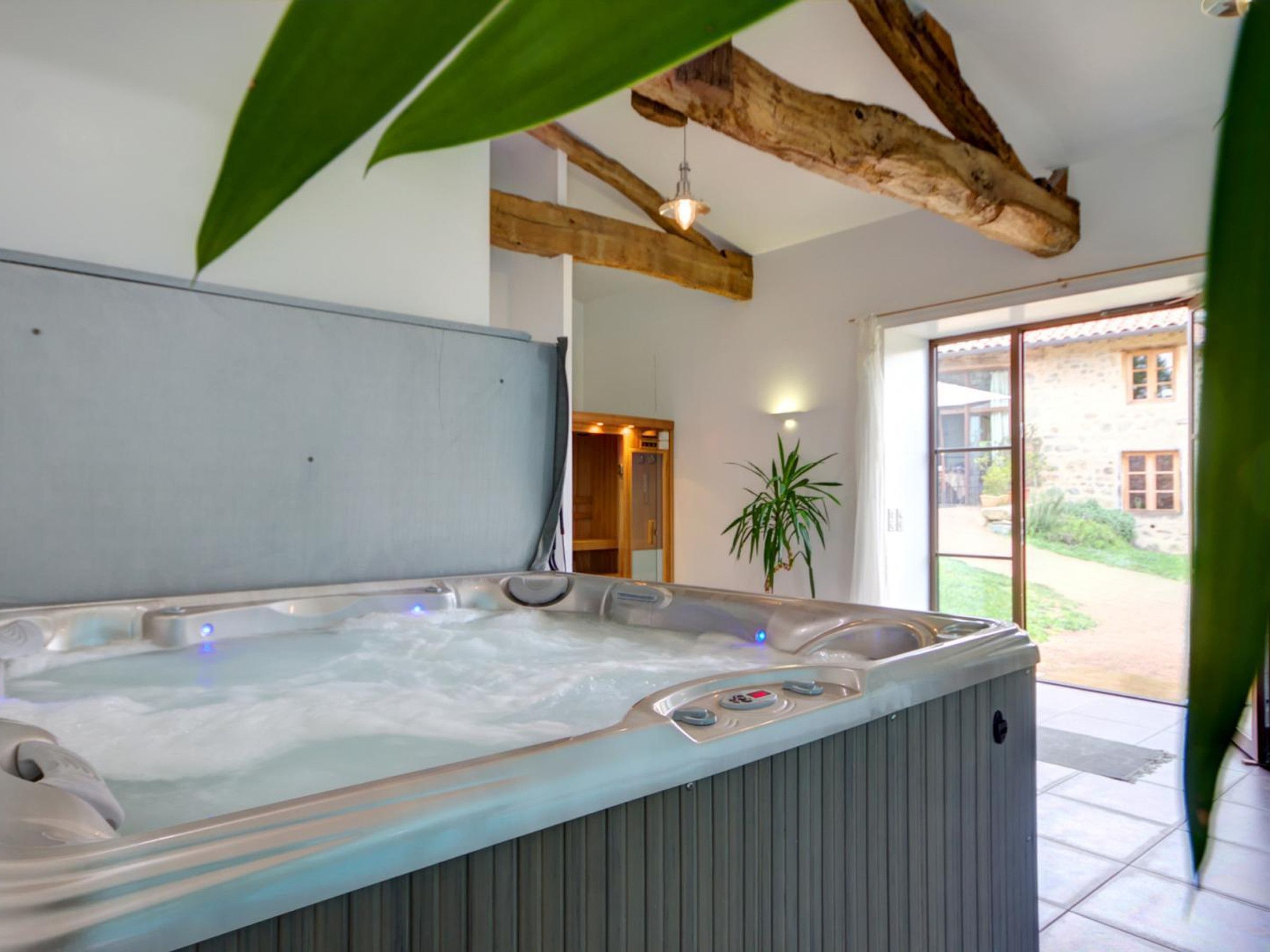 A hot tub with controls, a wooden ceiling beam, a potted plant, and a view of a stone building outside.