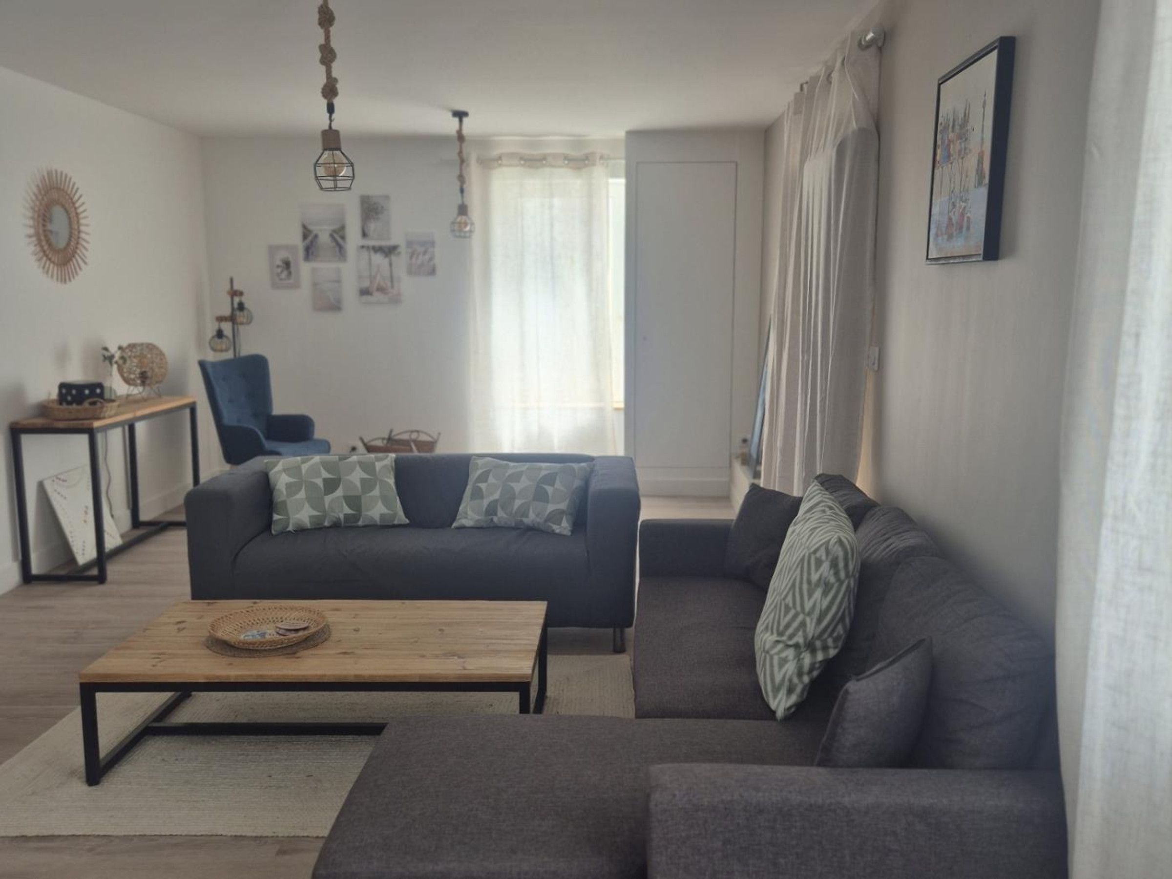 A living room with a sofa, a coffee table, and a chair. There are wall decorations and a window with curtains.