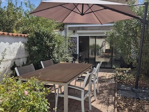 Un patio exterior con una mesa y sillas de madera, un paraguas grande y una puerta de cristal que da a una zona cubierta.