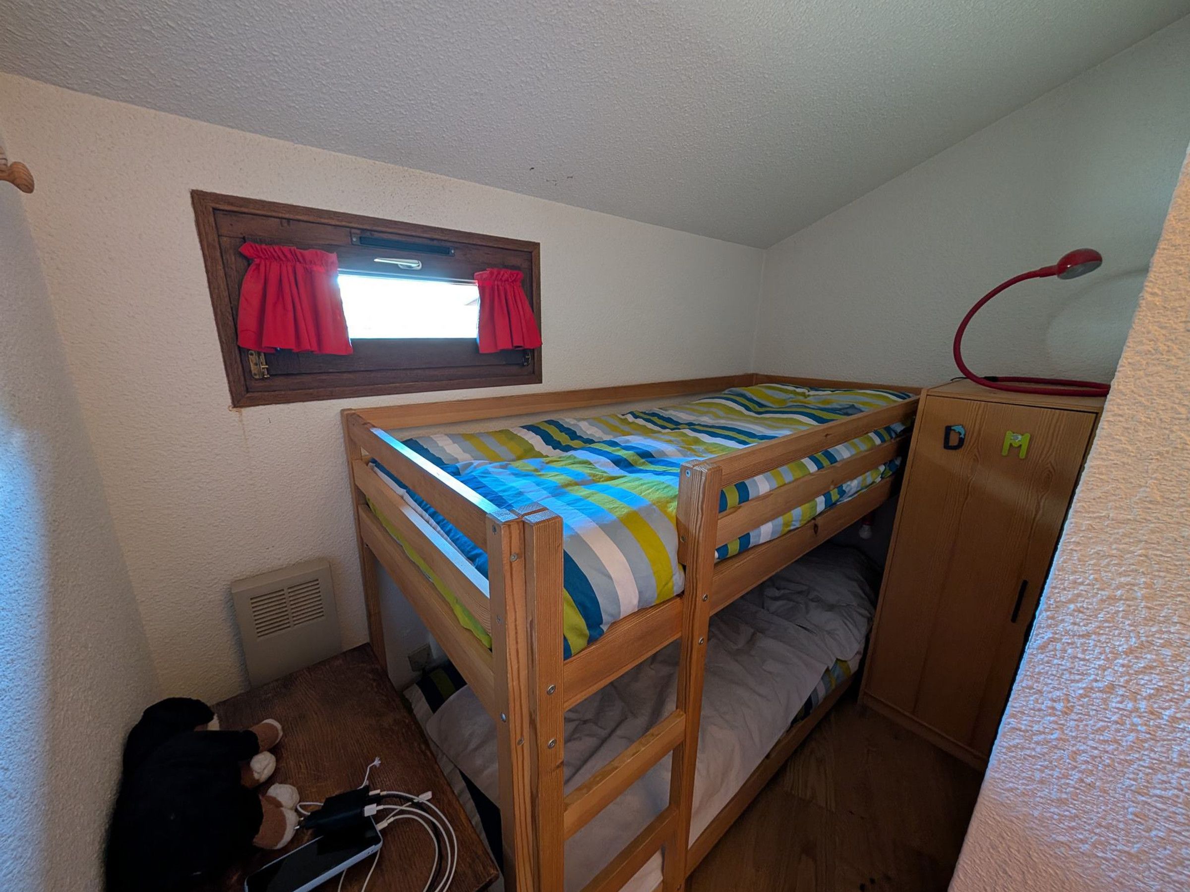 A bunk bed with a striped bedspread, a window with red curtains, a wooden wardrobe, and a desk with a computer and charger.