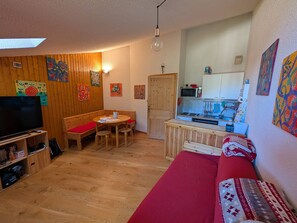 Ein gemütliches Zimmer mit Holzboden, einem roten Sofa, einer Holzbank, einem Fernseher und einer Küchenzeile mit Mikrowelle und Spüle.