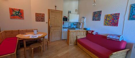 A compact living space with a wooden floor, a red sofa, a wooden dining table with chairs, a kitchenette, and various artworks on the walls.