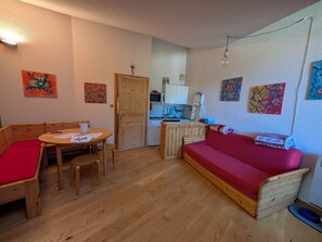 A compact living space with a wooden floor, a red sofa, a wooden dining table with chairs, a kitchenette, and various artworks on the walls. - Ski-in/ski-out duplex, pets welcome - 6 guests (Montgenèvre)