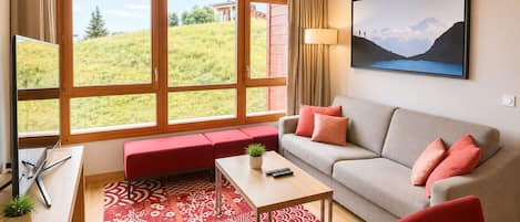 Un salon moderne avec un canapé, un banc rouge, une table basse et une grande fenêtre donnant sur un paysage verdoyant.
