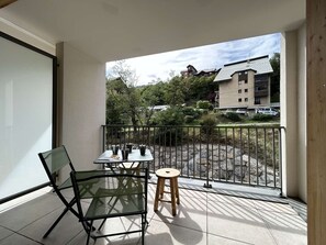 A balcony with a glass table and chairs, a small stool, and a view of a building and trees. - New and spacious apartment - 4 guests (Briançon)