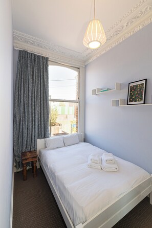 A bedroom with a bed, a window with curtains, a bedside table, a hanging light fixture, and a wall-mounted shelf with a framed picture. - Charming 2Bed Flat in the Heart of Brixton - Pass the Keys (London)