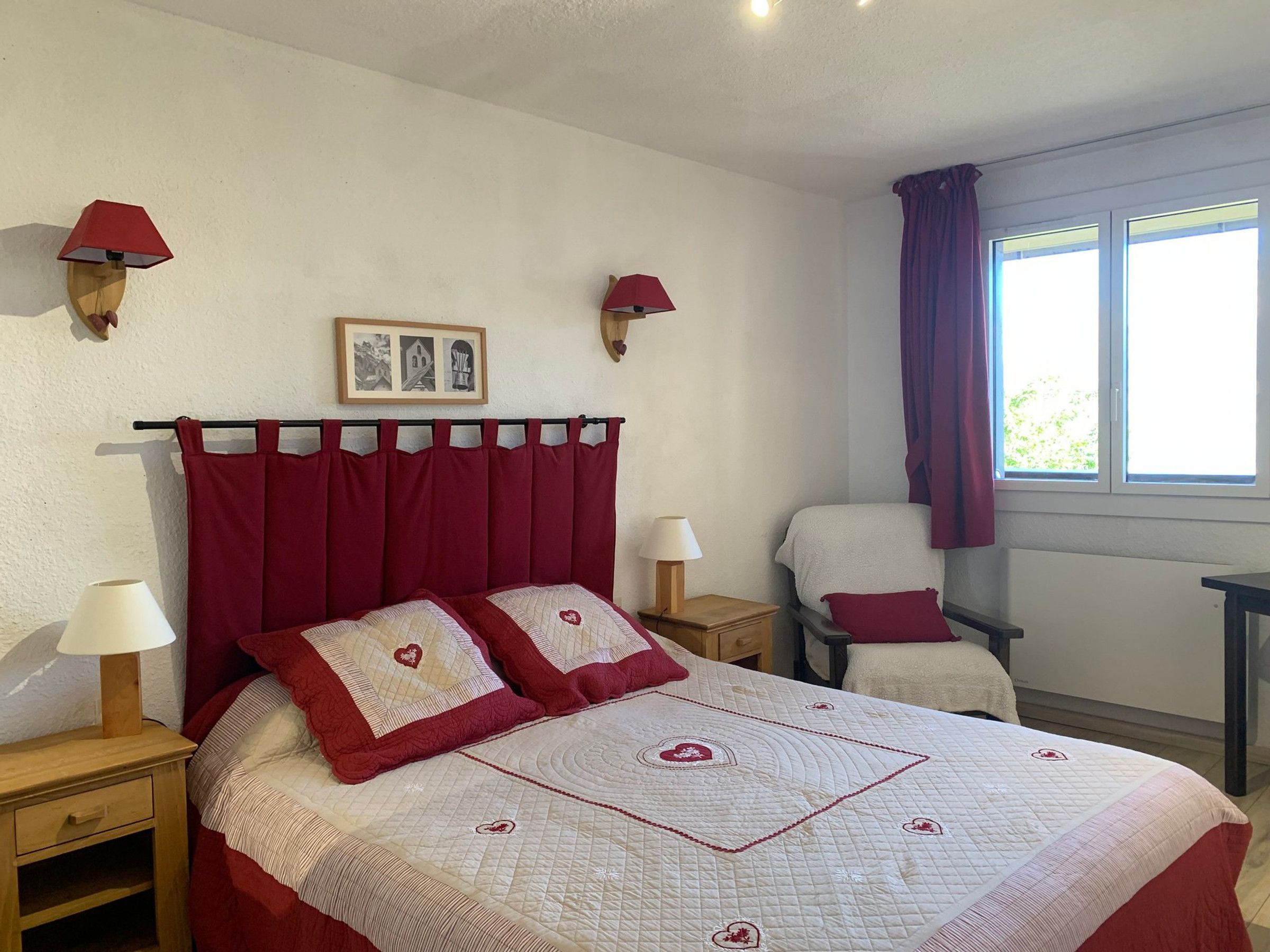 A bedroom with a bed, two bedside tables, a chair, a window with curtains, and wall-mounted lamps.