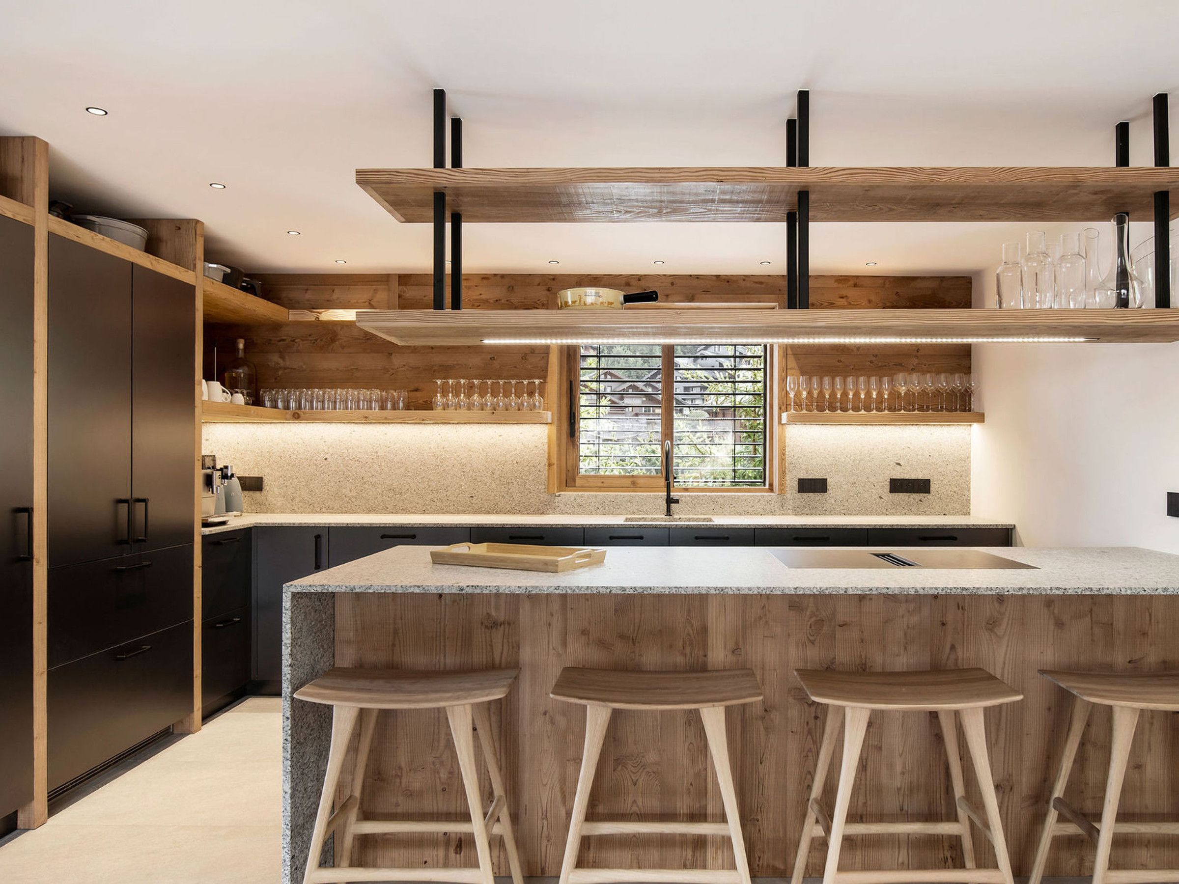 A modern kitchen with a central island, bar stools, wooden cabinetry, and a window with blinds.