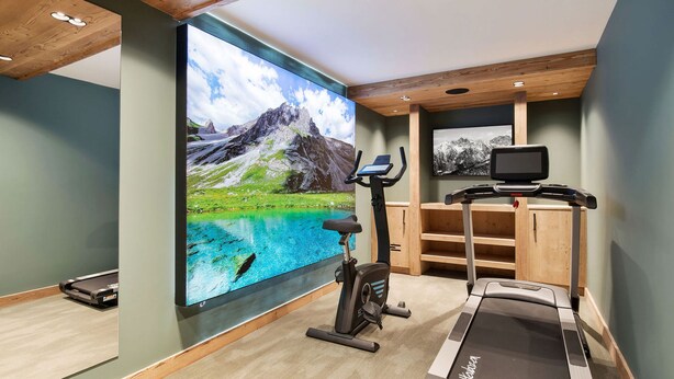 Une salle de sport à domicile équipée d’un tapis de course, d’un vélo d’appartement et d’un grand téléviseur fixé au mur, qui diffuse des images d’un paysage montagneux.