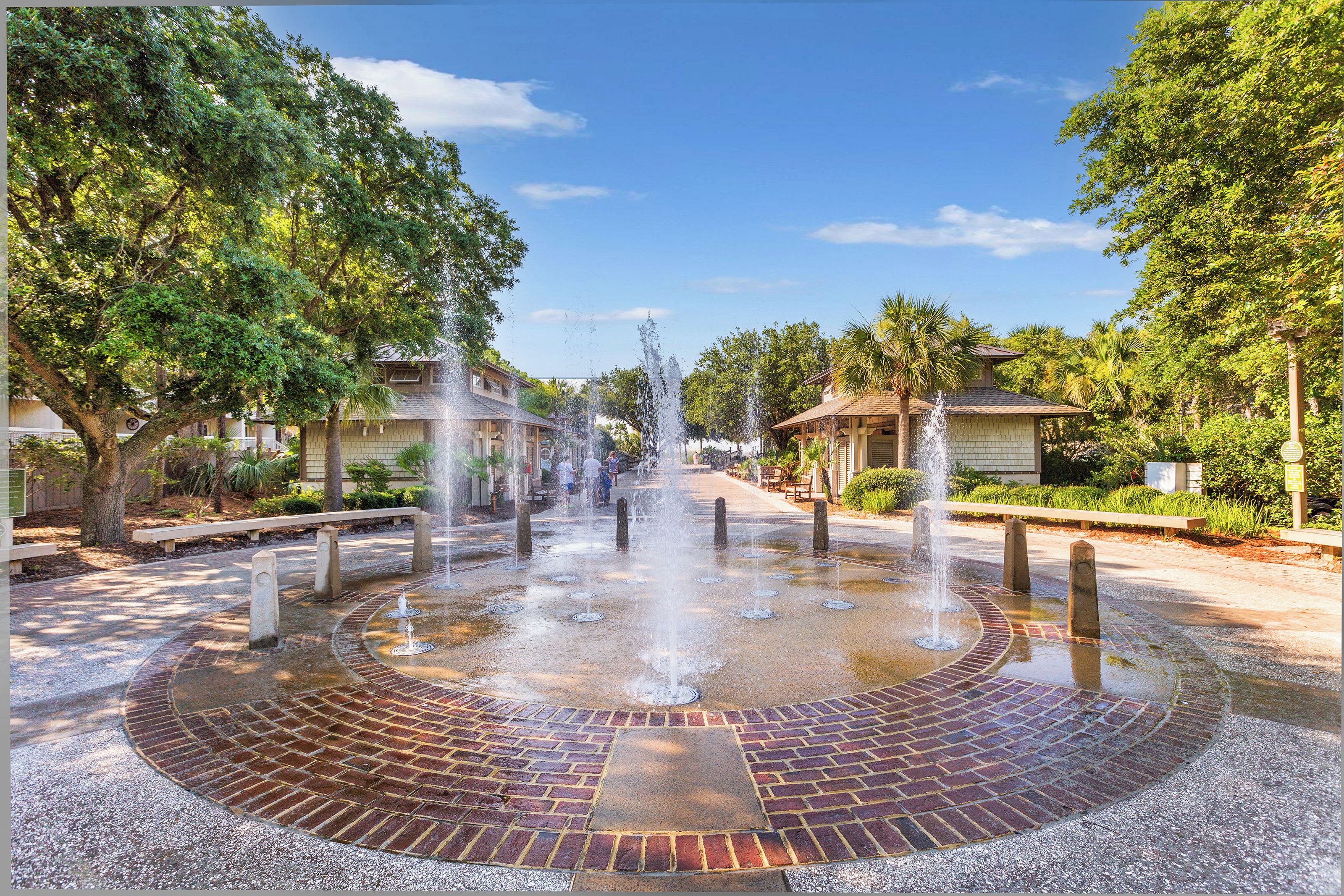 A circular brick fountain with water jets in a landscaped area with trees and buildings.