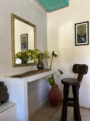 A room with a white wall, a mirror, a vase with flowers, a wooden chair, and a framed picture.