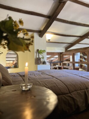 En seng med et beige quilt, et tændt stearinlys på et natbord og en vase med blomster.