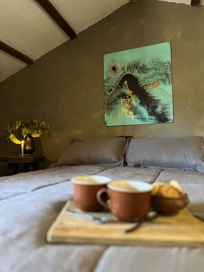Et soveværelse med seng, puder, et maleri, en vase med blomster og et fad med kopper og brød.