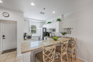 A kitchen with a central island, two bar stools, a refrigerator, a sink, and a window. - Olde Marco Inn Unit 208 | Walk To Food, Shops & More (Marco Island)