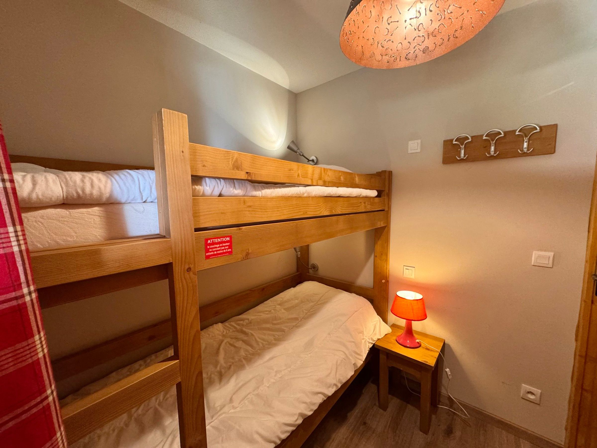 Une chambre avec des lits superposés, un panneau rouge, une petite table avec une lampe et un panneau mural avec des crochets.