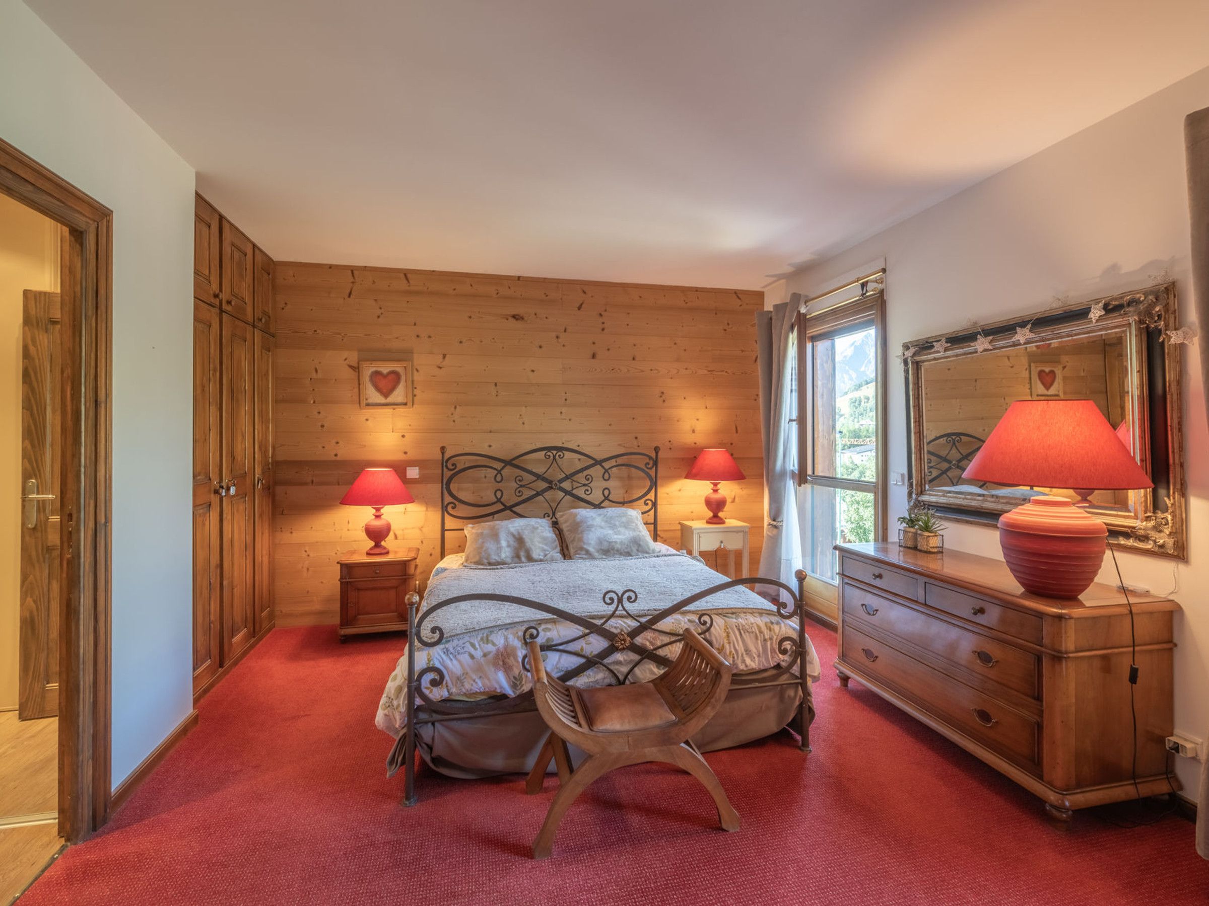 A bedroom with a wooden floor, a bed with a metal frame, two bedside tables with lamps, a dresser, and a large mirror on the wall.