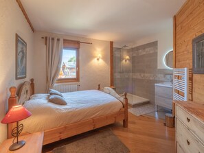 A bedroom with a wooden bed, a nightstand, a lamp, a window with curtains, a shower area, and a bathroom with a sink and mirror. - Mountain chalet, mountain view - 12 guests (Les Deux Alpes)