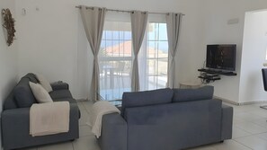 Ein modernes Wohnzimmer mit einem grauen Sofa, einem an der Wand befestigten Fernseher und einem großen Fenster mit Vorhängen.