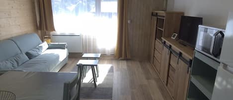 A compact living space with a sofa, a dining table, a kitchen area with a microwave, and a large window with curtains.